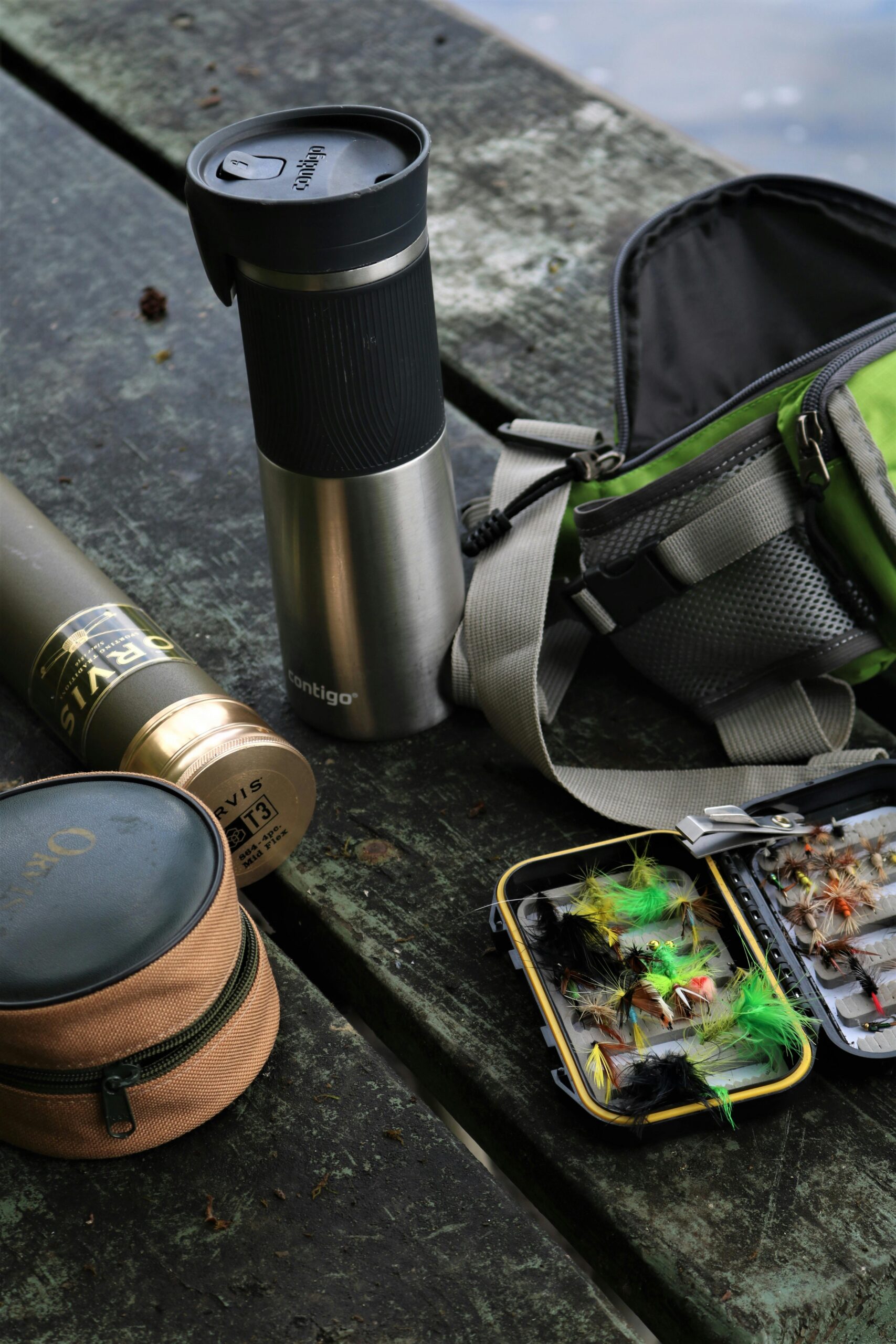 Fishing gear on a weathered wooden bench, including a Contigo thermos, rod tube, fly case with colorful artificial flies, and a green tackle bag